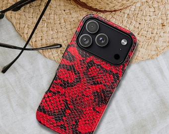 Puede incluir: Una funda de teléfono con estampado de piel de serpiente roja y negra. La funda está sobre un sombrero de paja y una superficie blanca. La funda del teléfono tiene un módulo de cámara negro con tres lentes y un flash. Gafas negras están al lado de la funda.