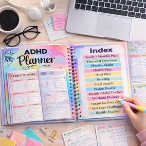 May include: An open ADHD planner with a spiral binding, featuring a colorful cover and index. The index lists sections like "Yearly + Monthly Plan" and "Health + Habit Tracker." A hand holds a pen, and a smartphone and laptop are also visible.