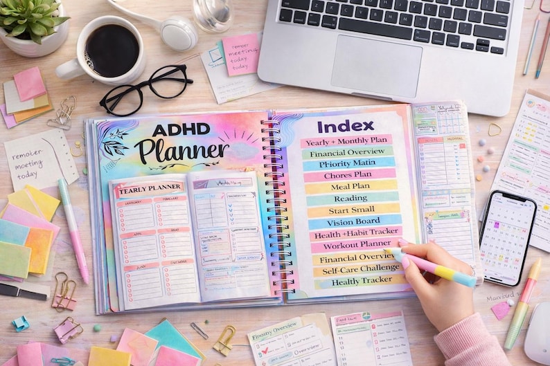 May include: An open ADHD planner with a spiral binding, featuring a colorful cover and index. The index lists sections like "Yearly + Monthly Plan" and "Health + Habit Tracker." A hand holds a pen, and a smartphone and laptop are also visible.