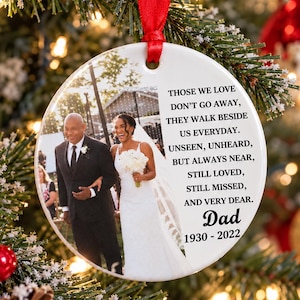 May include: A round white ceramic Christmas ornament hanging from a red ribbon. It features a printed photograph of a person in a suit walking with a person in a wedding dress, alongside the text: THOSE WE LOVE DON'T GO AWAY, THEY WALK BESIDE US EVERYDAY. UNSEEN, UNHEARD, BUT ALWAYS NEAR, STILL LOVED, STILL MISSED, AND VERY DEAR. Dad 1930 - 2022.