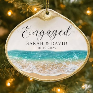 May include: A seashell-shaped Christmas ornament with a gold trim and a beach scene. The ornament reads "Engaged" above the names "SARAH & DAVID" and the date "10.19.2025". The ornament hangs from a gold string.