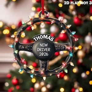 Pode incluir: Um ornamento plano 2D em forma de volante preto pendurado por uma fita vermelha. O volante est&aacute; envolto em luzes de Natal coloridas e apresenta o texto THOMAS, NEW DRIVER, 2026 impresso em branco no centro. O texto 2D FLAT NOT 3D &eacute; vis&iacute;vel.