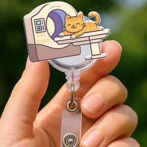 May include: A retractable badge reel featuring a cartoon cat undergoing a CAT SCAN. The cat is orange with a smiling face. The badge reel has a clear plastic strap and a metal clip. The text "CAT SCAN" is visible on the machine.