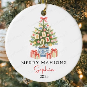 May include: White ceramic Christmas ornament with a festive design. It features a Christmas tree decorated with Mahjong tiles, a red bow, and gift boxes. The text "MERRY MAHJONG Sophia 2025" is printed on the ornament.