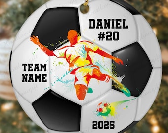 Pode incluir: Enfeite de Natal em forma de bola de futebol branca e preta, com um cordão dourado. Apresenta uma ilustração colorida de um jogador de futebol. O texto inclui "DANIEL #20", "TEAM NAME" e "2025".