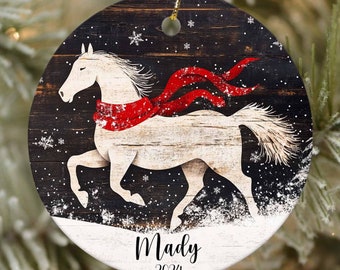 Puede incluir: Un caballo blanco con una bufanda roja corre a través de una escena de bosque nevado. El adorno es redondo y tiene un fondo de madera oscura con copos de nieve blancos. El texto "Mady 2024" está impreso en la parte inferior del adorno.