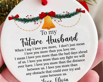 May include: White ceramic Christmas ornament with a red ribbon and a gold bell. The ornament has a message that reads "To my Future Husband When I say I love you more, I don't just mean I love you more than you love me. I mean I love you more than the bad days ahead of us. I love you more than any fight we will ever have. I love you more than the distance between us. I love you more than any obstacle that could ever try and come between us. Love, Olivia"