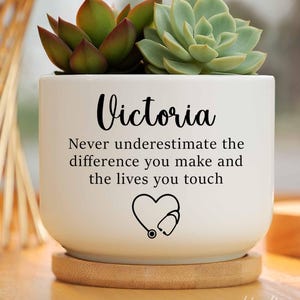 May include: White ceramic planter with the name "Victoria" and the text "Never underestimate the difference you make and the lives you touch". A heart with a stethoscope is below the text. The planter has two succulents inside, one green and one red.