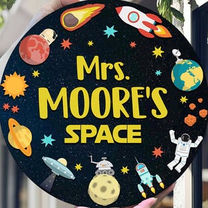 May include: A round, black sign with a space theme, featuring planets, stars, a rocket, and an astronaut. The text "Mrs. MOORE'S SPACE" is in yellow. The sign is held up by a person.