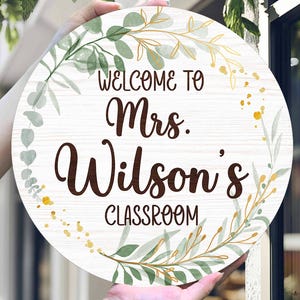 May include: A round wooden sign with the text "WELCOME TO Mrs. Wilson's CLASSROOM" in brown script. The sign is decorated with green and gold botanical illustrations and is held by two hands.
