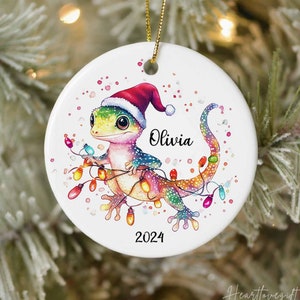 May include: A white ceramic Christmas ornament featuring a cartoon gecko wearing a Santa hat and holding colorful Christmas lights. The ornament is personalized with the name "Olivia" and the year "2024".