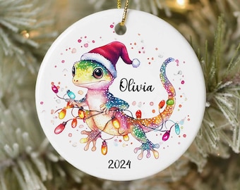 Può includere: Un ornamento natalizio in ceramica bianca con un geco dei cartoni animati che indossa un cappello di Babbo Natale e tiene delle luci natalizie colorate. L'ornamento è personalizzato con il nome "Olivia" e l'anno "2024".