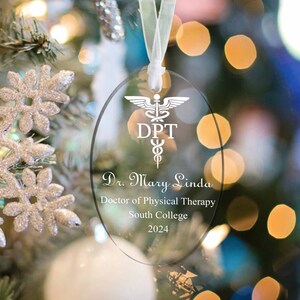 May include: A clear oval glass ornament with a white ribbon. The ornament has a white caduceus symbol with the letters "DPT" and the text "Dr. Mary Linda, Doctor of Physical Therapy, South College, 2024".