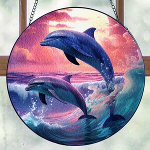 May include: A round stained glass suncatcher featuring two dolphins jumping out of the water against a colorful sunset background. The dolphins are blue and gray, and the water is a turquoise blue. The suncatcher is perfect for adding a touch of whimsy to any window.