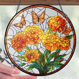 May include: Round stained glass window art featuring orange marigold flowers and butterflies. The artwork is framed with a dark border and hangs from a chain. The flowers are in full bloom, with green leaves and stems.