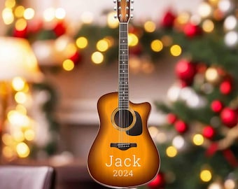May include: A brown acoustic guitar ornament with the name "Jack" and the year "2024" engraved on it. The ornament is hanging from a red ribbon in front of a blurred background of a Christmas tree with lights and ornaments.