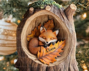 May include: A Christmas tree ornament featuring a sleeping fox nestled in a hollowed-out tree trunk. The fox is orange and white, resting on autumn leaves. The ornament has a red ribbon for hanging and the name "Olivia" is engraved on a leaf.