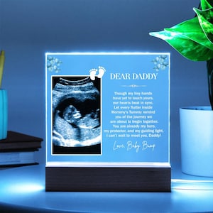 May include: A blue light-up photo frame with a sonogram image and a message to "Dear Daddy" that reads "Though my tiny hands have yet to touch yours, our hearts beat in sync. Let every flutter inside Mommy's Tummy remind you of the journey we are about to begin together. You are already my hero, my protector, and my guiding light. I can't wait to meet you, Daddy! Love, Baby Bump"
