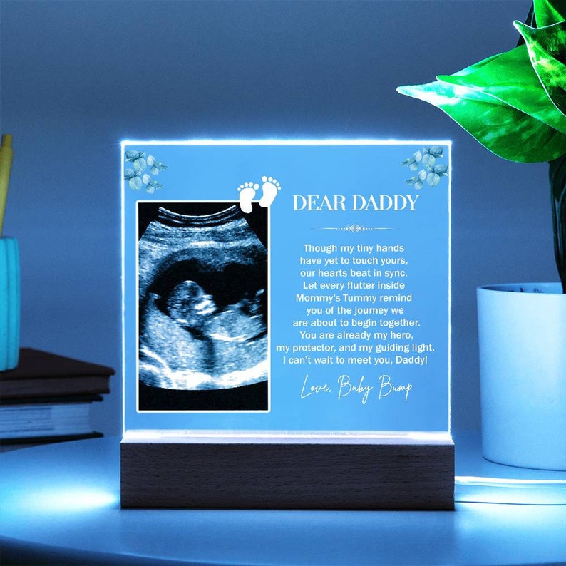 May include: A blue light-up photo frame with a sonogram image and a message to "Dear Daddy" that reads "Though my tiny hands have yet to touch yours, our hearts beat in sync. Let every flutter inside Mommy's Tummy remind you of the journey we are about to begin together. You are already my hero, my protector, and my guiding light. I can't wait to meet you, Daddy! Love, Baby Bump"