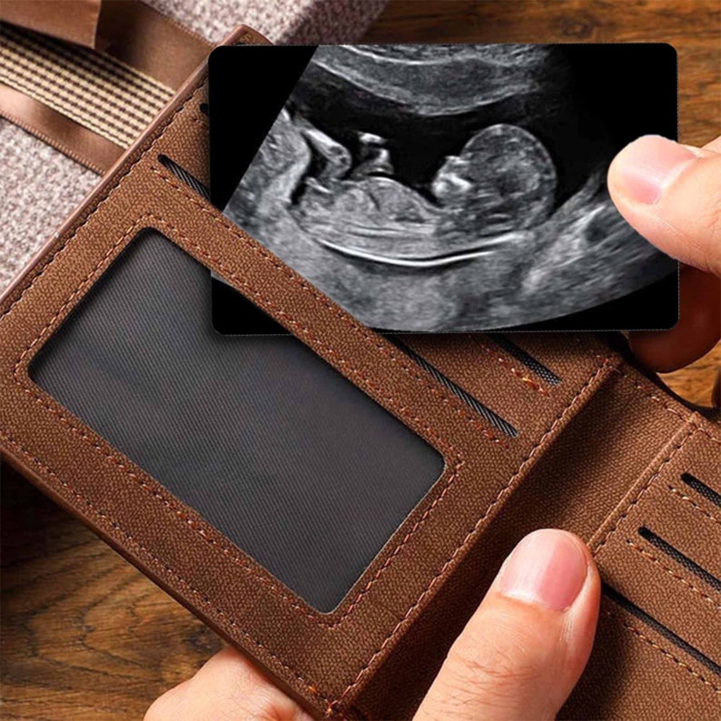 May include: A brown leather wallet with a clear plastic window for displaying a photo or card. The wallet is open and a black and white ultrasound image of a baby is visible in the window.