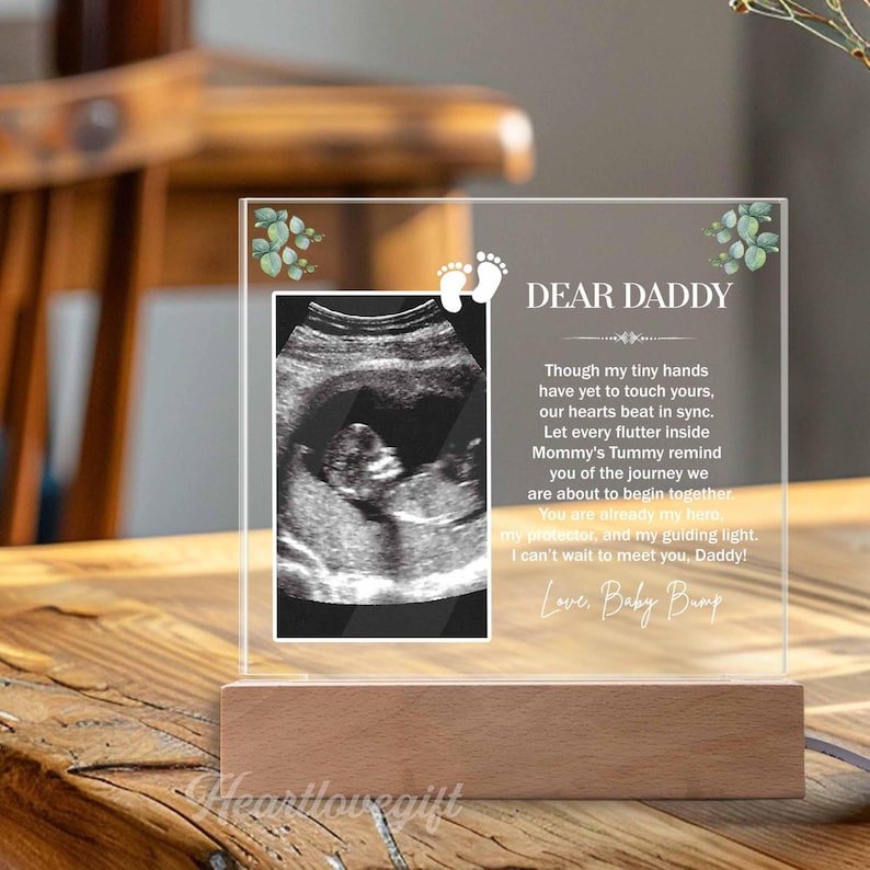 May include: A clear acrylic frame with a wooden base, displaying a black and white ultrasound image and a message to "Dear Daddy". The message reads, "Though my tiny hands have yet to touch yours, our hearts beat in sync. Let every flutter inside Mommy's Tummy remind you of the journey we are about to begin together. You are already my hero, my protector, and my guiding light. I can't wait to meet you, Daddy! Love, Baby Bump."
