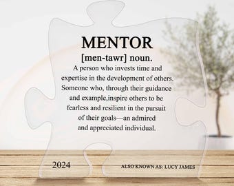 May include: A clear acrylic puzzle piece with the word "MENTOR" in bold black letters and a definition of the word below. The text reads "[men-tawr] noun. A person who invests time and expertise in the development of others. Someone who, through their guidance and example, inspire others to be fearless and resilient in the pursuit of their goals - an admired and appreciated individual." The year 2024 and the text "ALSO KNOWN AS: LUCY JAMES" are printed below the definition.