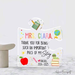 May include: A clear acrylic puzzle piece with colorful illustrations and text. The text reads "MRS. CLARA... THANK YOU FOR BEING SUCH AN IMPORTANT PIECE OF MY Story." Includes a red apple, pencil, globe, and a worm with glasses.