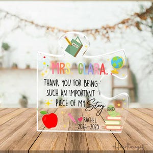 May include: A clear acrylic puzzle piece with the text "MRS. CLARA. THANK YOU FOR BEING SUCH AN IMPORTANT PIECE OF MY Story." Includes a red apple, pencil, globe, and a worm on books. The name "RACHEL 2024-2025" is also included.