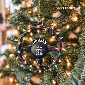Pode incluir: Um ornamento de volante plano e bidimensional com uma corrente de luzes de Natal coloridas enroladas na borda. O centro exibe o texto 'THOMAS', 'NEW DRIVER' e '2026'. O texto '2D FLAT NOT 3D' &eacute; vis&iacute;vel no topo.
