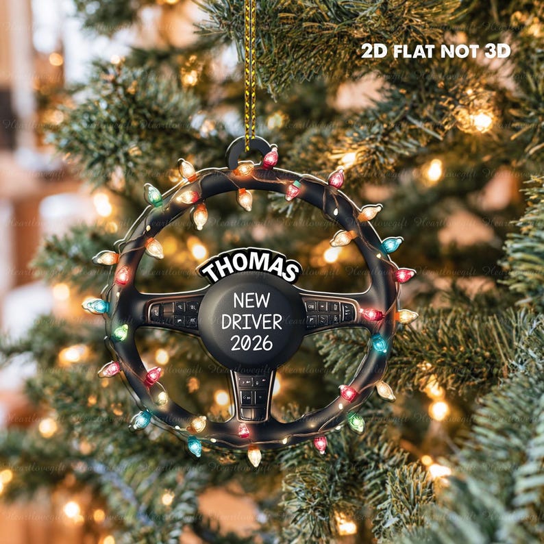 Pode incluir: Um ornamento de volante plano e bidimensional com uma corrente de luzes de Natal coloridas enroladas na borda. O centro exibe o texto 'THOMAS', 'NEW DRIVER' e '2026'. O texto '2D FLAT NOT 3D' &eacute; vis&iacute;vel no topo.