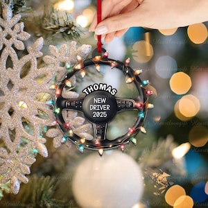 Pode incluir: Um ornamento de Natal em forma de volante preto circular decorado com luzes coloridas. O centro apresenta o texto "THOMAS" no topo e "NEW DRIVER 2025" abaixo. O ornamento est&aacute; pendurado por uma fita vermelha num fundo festivo.