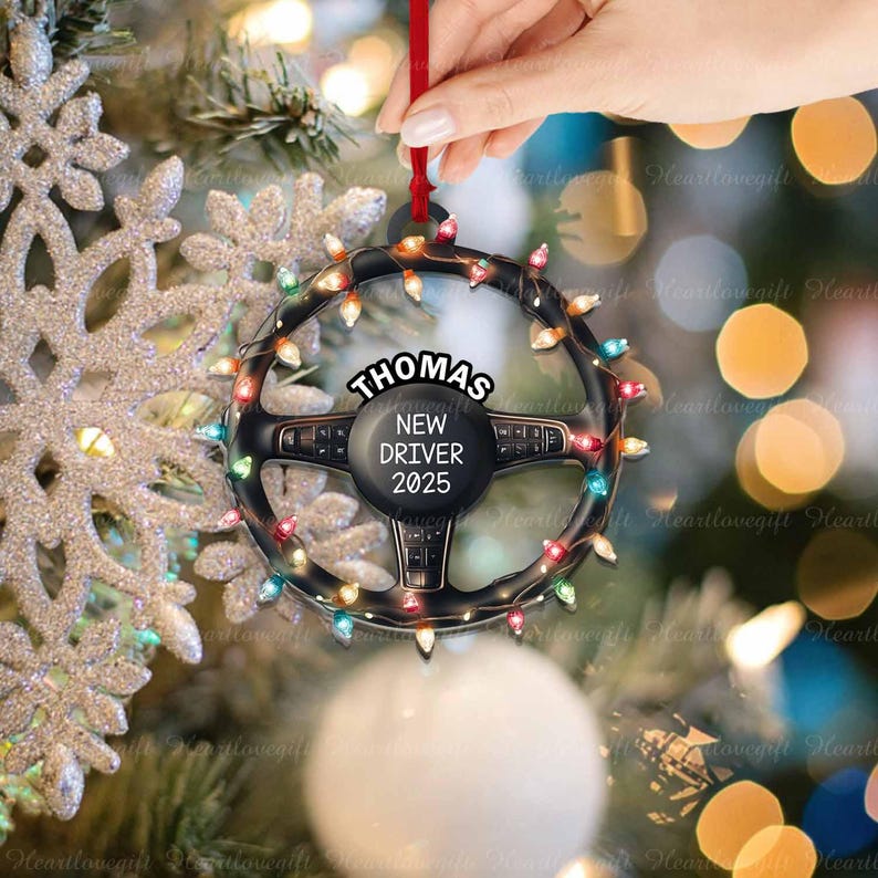 Pode incluir: Um ornamento de Natal em forma de volante preto circular decorado com luzes coloridas. O centro apresenta o texto "THOMAS" no topo e "NEW DRIVER 2025" abaixo. O ornamento est&aacute; pendurado por uma fita vermelha num fundo festivo.