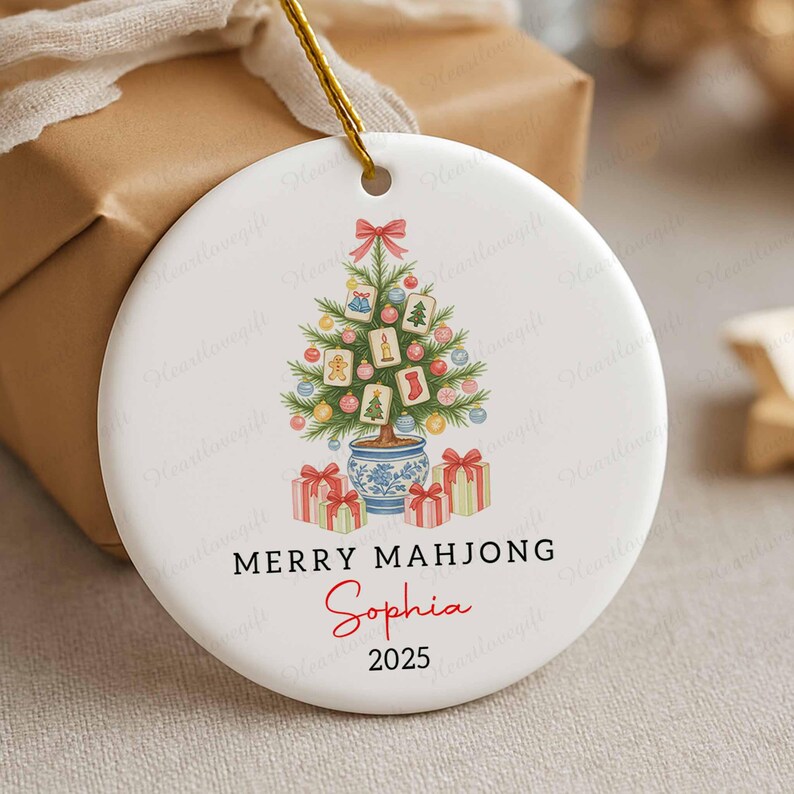 May include: White ceramic Christmas ornament with a festive Mahjong-themed design. The ornament features a Christmas tree decorated with Mahjong tiles, ornaments, and gifts. Text reads "MERRY MAHJONG Sophia 2025".