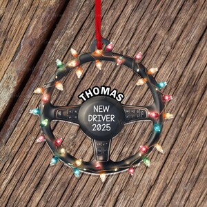 Pode incluir: Um ornamento em forma de volante preto decorado com luzes de Natal multicoloridas. O centro apresenta o texto THOMAS NEW DRIVER 2025 em letras brancas. Uma fita vermelha est&aacute; presa ao topo para pendurar este enfeite circular.