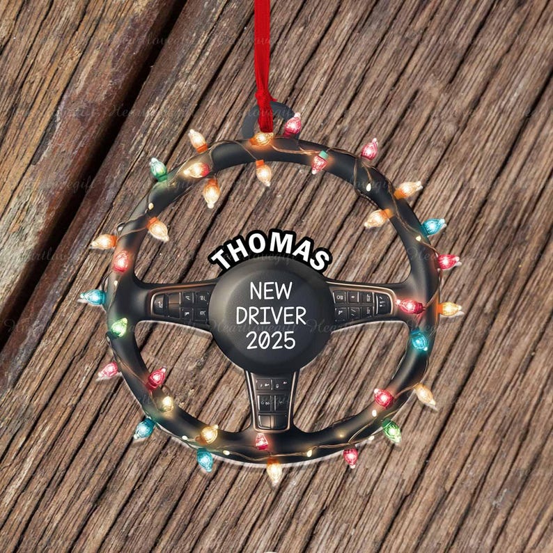 Pode incluir: Um ornamento em forma de volante preto decorado com luzes de Natal multicoloridas. O centro apresenta o texto THOMAS NEW DRIVER 2025 em letras brancas. Uma fita vermelha est&aacute; presa ao topo para pendurar este enfeite circular.
