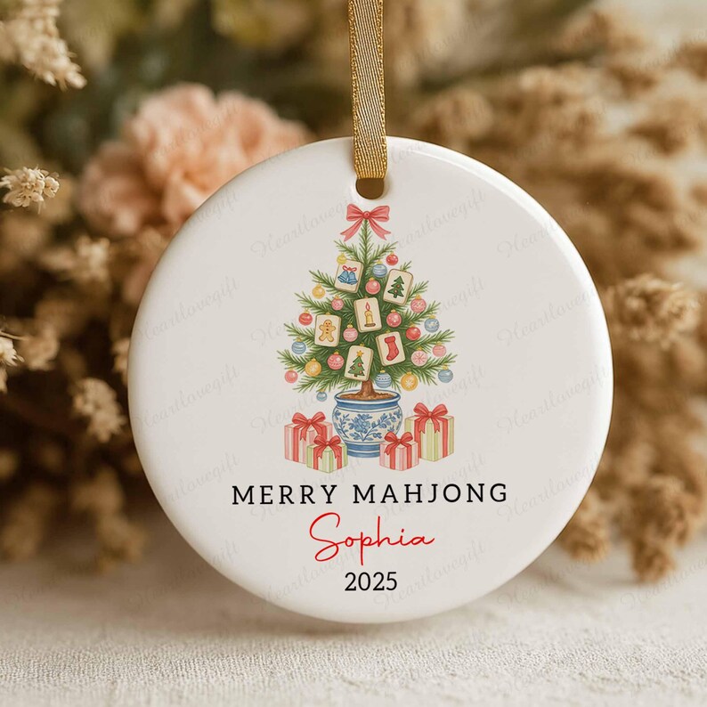 May include: A white ceramic Christmas ornament with a gold ribbon. The ornament features a Christmas tree design with Mahjong tiles, gifts, and the text "MERRY MAHJONG Sophia 2025".