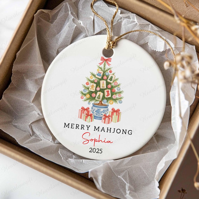 May include: A white ceramic Christmas ornament with a festive design. It features a Christmas tree decorated with Mahjong tiles, ornaments, and gift boxes. The text reads "MERRY MAHJONG" and "Sophia 2025". A gold string is attached for hanging.