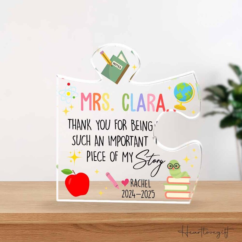 May include: A clear acrylic puzzle piece with the text "MRS. CLARA. THANK YOU FOR BEING SUCH AN IMPORTANT PIECE OF MY Story RACHEL 2024-2025". The piece has colorful illustrations of a notebook, globe, apple, pencil, and a worm on books.