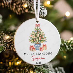 May include: A white ceramic Christmas ornament with a festive design. It features a Christmas tree decorated with ornaments and gifts. The text "MERRY MAHJONG" and the name "Sophia" are printed on the ornament, along with the year 2025.