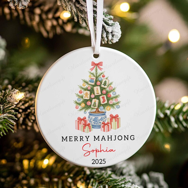 May include: A white ceramic Christmas ornament with a festive design. It features a Christmas tree decorated with ornaments and gifts. The text "MERRY MAHJONG" and the name "Sophia" are printed on the ornament, along with the year 2025.