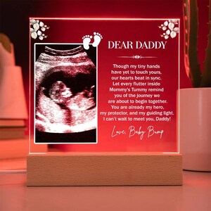 May include: A red illuminated acrylic plaque with a white wooden base. The plaque features a black and white ultrasound image of a baby in the womb. The text on the plaque reads "DEAR DADDY Though my tiny hands have yet to touch yours, our hearts beat in sync. Let every flutter inside Mommy's Tummy remind you of the journey we are about to begin together. You are already my hero, my protector, and my guiding light. I can't wait to meet you, Daddy! Love, Baby Bump"