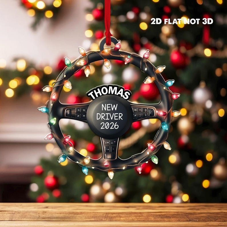Pode incluir: Um ornamento plano 2D em forma de volante preto pendurado por uma fita vermelha. O volante est&aacute; envolto em luzes de Natal coloridas e apresenta o texto THOMAS, NEW DRIVER, 2026 impresso em branco no centro. O texto 2D FLAT NOT 3D &eacute; vis&iacute;vel.