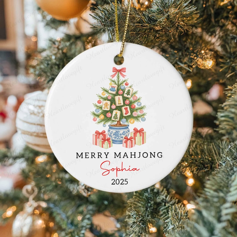 May include: White ceramic Christmas ornament with a festive design. It features a Christmas tree decorated with Mahjong tiles, a red bow, and gift boxes. The text "MERRY MAHJONG Sophia 2025" is printed on the ornament.