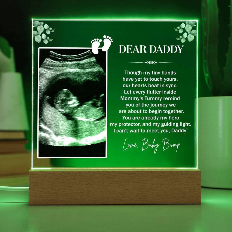 May include: A green illuminated acrylic plaque with a sonogram image and a message to "Dear Daddy" from the "Baby Bump". The message reads, "Though my tiny hands have yet to touch yours, our hearts beat in sync. Let every flutter inside Mommy's Tummy remind you of the journey we are about to begin together. You are already my hero, my protector, and my guiding light. I can't wait to meet you, Daddy! Love, Baby Bump."