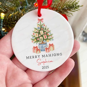 May include: A white ceramic Christmas ornament with a red ribbon. The ornament features a Christmas tree design with Mahjong tiles, gift boxes, and the text "MERRY MAHJONG Sophia 2025".