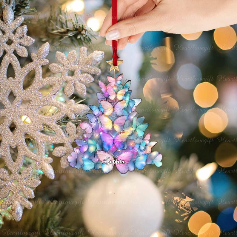 May include: A Christmas tree ornament shaped like a tree, made of colorful butterflies. The ornament is held by a red ribbon with a gold star. The name "Sophia" is written on the ornament.