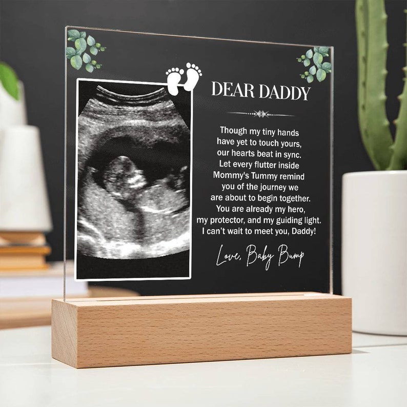 May include: A clear acrylic photo frame with a wooden base, displaying a black and white ultrasound image and a message to "Dear Daddy". The message reads, "Though my tiny hands have yet to touch yours, our hearts beat in sync. Let every flutter inside Mommy's Tummy remind you of the journey we are about to begin together. You are already my hero, my protector, and my guiding light. I can't wait to meet you, Daddy! Love, Baby Bump."