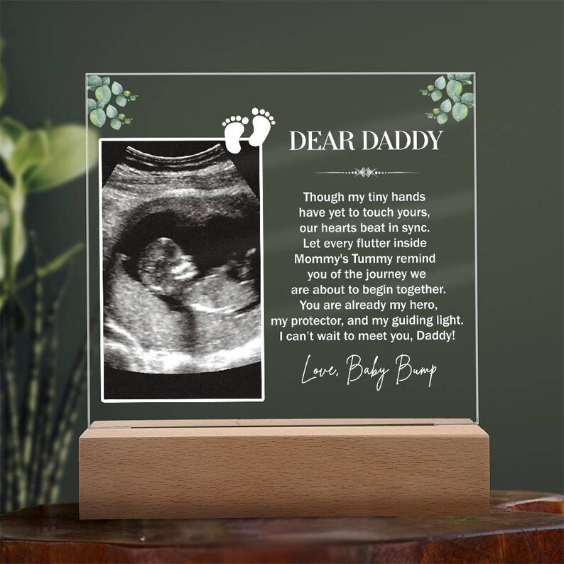 May include: A clear acrylic plaque with a wooden base, featuring a sonogram image and a heartfelt message to a father-to-be. The message reads "Dear Daddy, Though my tiny hands have yet to touch yours, our hearts beat in sync. Let every flutter inside Mommy's Tummy remind you of the journey we are about to begin together. You are already my hero, my protector, and my guiding light. I can't wait to meet you, Daddy! Love, Baby Bump."