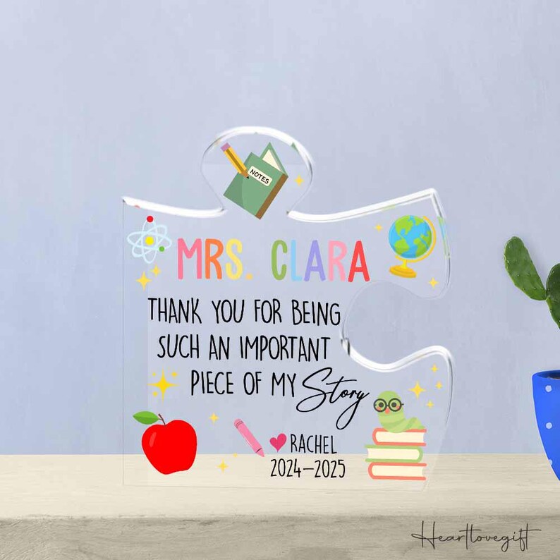 May include: Clear acrylic puzzle piece with the text "MRS. CLARA THANK YOU FOR BEING SUCH AN IMPORTANT PIECE OF MY Story RACHEL 2024-2025". Features a red apple, pencil, globe, and a worm with books.
