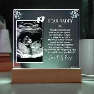 May include: A white acrylic plaque with a black and white ultrasound image and a message to "Dear Daddy" from "Baby Bump". The message reads, "Though my tiny hands have yet to touch yours, our hearts beat in sync. Let every flutter inside Mommy's Tummy remind you of the journey we are about to begin together. You are already my hero, my protector, and my guiding light. I can't wait to meet you, Daddy!"